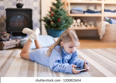 Child Is Using Mobile Phone Under Christmas Tree At Home. Kid Is Playing In Children Games, Looking At Devices. Girl In Blue Knitted Sweater With Her Present Or Gift Is Laying On Warm Floor.