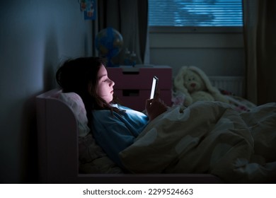 A child using smart phone lying in bed late at night, playing games, watching videos online, scrolling screen. Children's screen addiction. Child's room at night. 