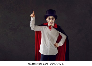 Child In Vampire Costume Against Dark Studio Background. Kid Dressed As Dracula For Halloween Party. Boy Wearing Top Hat, Red And Black Cape, White Shirt, Fake Fangs And Scary Makeup Points Finger Up