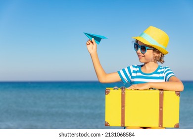 Child With Vintage Suitcase On Summer Vacation. Girl Having Fun On The Beach. Kid With Paper Airplane Sitting Against Sea And Sky Background.  Travel And Adventure Concept