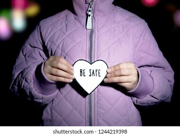 Child In Violet Quilted Jacket Holding Heart-shaped Pedestrian Safety Reflector With Text Be Safe Outdoors With Dark Background And Bokeh City Street Lights