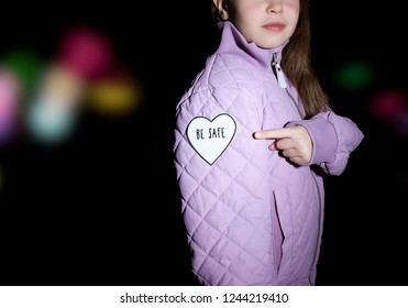 Child In Violet Quilted Jacket Pointing At Heart-shaped Pedestrian Safety Reflector With Text Be Safe Outdoors With Dark Background And Bokeh City Street Lights