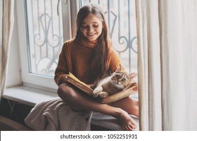 Child In Warm Woolen Sweater Seating On Window Sill And Reading A Book. Winter Weekend With Cat At Home. Cozy Scene, Hygge Concept.