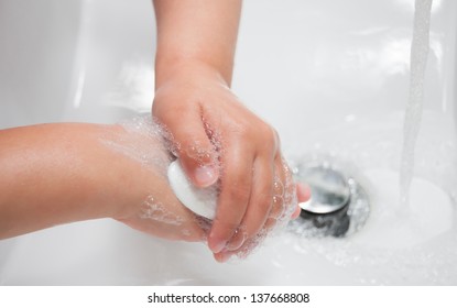 Child Washing Hands In A White Basin With A Bar Of White Soap.