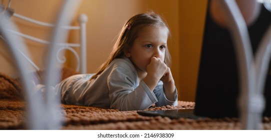 Child Watching Cartoons On Laptop. Serious Kid With Technology At Home On Bed Spending Time On-line. Education And Entertainment For Toddlers. Girl In Cozy Atmospheric Room Over The Window