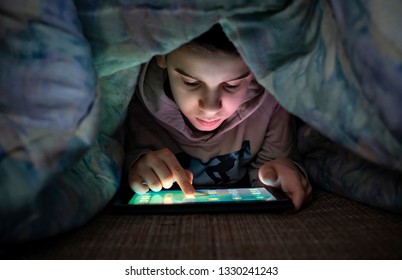 Child Watching His Tablet In The Bed. Illuminated Child Face From Device Screen. Boy Under The Covers Hold A Tablet. Night Time.