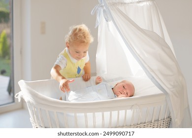 Child watching newborn baby in bassinet. Sibling meeting new family member. Kids play in white bedroom. Nursery crib bedding and textile for young kid. Siblings love. - Powered by Shutterstock