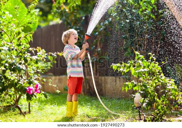 Child watering flowers and plants in garden. Kid\
with water hose in sunny blooming backyard. Little boy gardening.\
Summer fun outdoor at home. Family activity in spring. Children\
help parents.