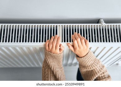 Child wearing brown woolly jacket warming cold hand in front of heating radiator in winter time. Electric or gas heater at home. Heating season, cold room, heating problems.