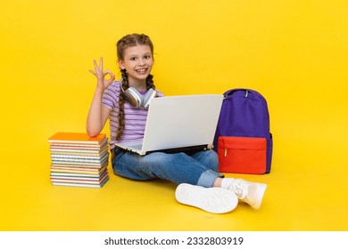 A child wearing headphones with a laptop shows the OK sign. A young girl with books and a backpack does homework online with a teacher. Yellow isolated background. - Powered by Shutterstock