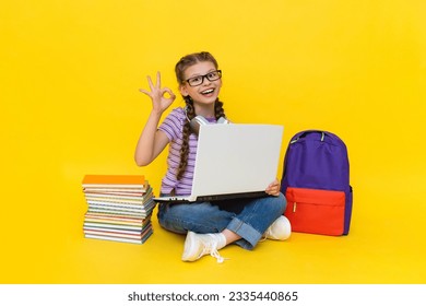 A child wearing headphones with a laptop shows the OK sign. A young girl with books and a backpack does homework online with a teacher. Yellow isolated background. - Powered by Shutterstock