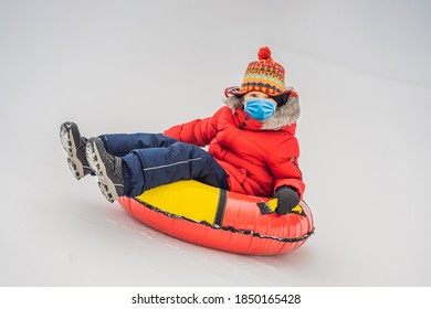 Child Wearing A Medical Mask During COVID-19 Coronavirus Having Fun On Snow Tube. Boy Is Riding A Tubing. Winter Fun For Children