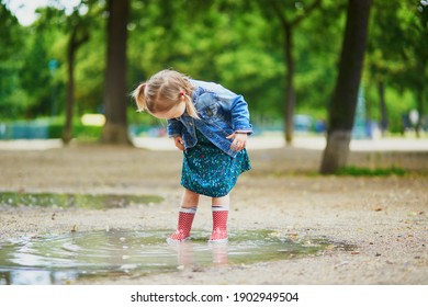 Child wearing red rain boots and jumping in puddle on a summer day. Adorable toddler girl having fun with water and mud in park on a rainy day. Outdoor activities for kids