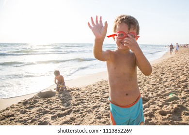 Child Wearing Swimming Goggles Say Hello With His Hand By The Sea