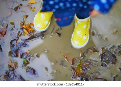 Child wearing yellow rain boots and jumping in dirty puddle on a fall day. Toddler girl having fun with water and mud in park on a rainy day. Outdoor autumn activities for kids