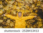 A child wearing a yellow raincoat lies happily among a in golden autumn leaves, smiling and enjoying the crisp fall weather. The leaves create a vibrant backdrop, showcasing the beauty of the season