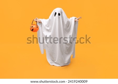 Child in white ghost costume holding pumpkin bucket on orange background. Halloween celebration