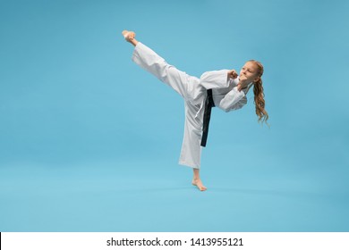 Child In White Kimono With Black Belt, Barefoot Posing At Camera On Blue Background, Looking Away. Karate Girl Training Pose Kick Foot Forward. Japanese Arts Of Health And Defense.