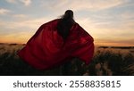 Child winner runs in superhero costume in nature park. Kid girl game concept. Little girl in red mantle play superhero against sun sky. Happy child runs through green meadow wearing red superhero cape