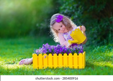 Child Working In The Garden. Kids Gardening. Children Watering Flowers. Little Girl With Water Can On A Green Lawn In The Backyard In Summer. Toddler Kid Playing Outdoors Planting Purple Flower Pots.
