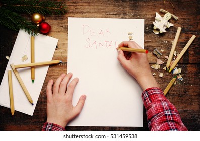 Child Writes The Letter To Santa Claus. Child's Hands, The Sheet Of Paper, Pencils And Christmas Decorations On A Wooden Surface. Concept Of New Year And Christmas. Top View