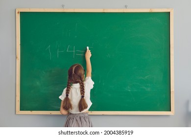 Child Writing On Chalkboard During Math Class. Back View Of Little Student Girl Standing In Front Of Green School Board, Holding Piece Of Chalk And Writing Down A Simple Elementary Arithmetic Sum