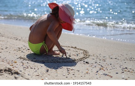 Child Writing With A Stick On The Sand. Girl With Hat In Summer On The Beach Relaxing And Playing.
