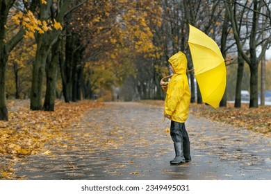 Child in yellow raincoat, rubber boots and an umbrella in hands is walking in the autumn park.