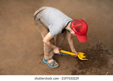 A Child With A Yellow Shovel Digs The Sand. The Boy Is Picking In The Mud. The Preschooler Has Stained His Clothes. The Hyperactive Boy Is Walking In The Yard.