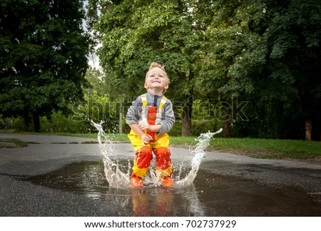 Child in a yellow waterproof coat in the rain jumping in puddles. A boy stands with his head bowed in rain, a bright raincoat. Kid playing autumn park. Outdoor fun by any weather