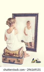 Child Or Young Girl Fixing Her Hair While Looking At Herself In A Mirror, Sitting On Vintage Luggage, With A Fish Tail Braid In Her Hair. Vintage Or Retro Filter Applied.