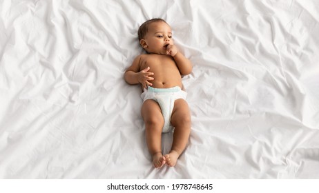 Childcare Concept. Above top high angle view portrait of cute little African American baby wearing diaper lying on the white blanket sheets at home with finger in mouth. Black infant child resting
