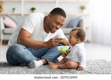 Childcare Concept. Caring Young Black Father Feeding His Adorable Baby Son With Spoon At Home, Loving African American Daddy Giving Food To Little Infant Child While Sitting Together In Living Room