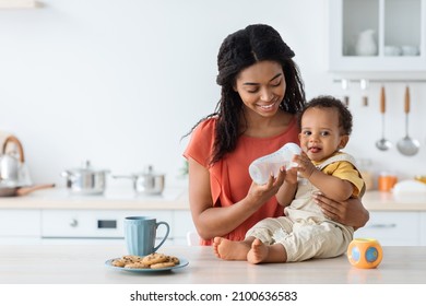 Childcare Concept. Happy Black Mom Giving Water Bottle To Cute Toddler Son While Spending Time Together In Kitchen, Adorable Infant Baby Enjoying Healthy Drink While Sitting On Counter Table