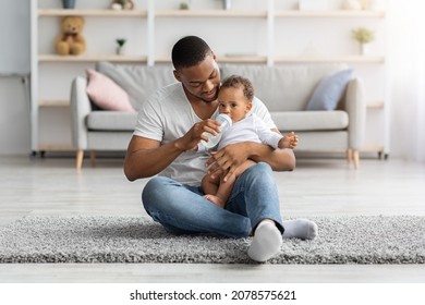 Childcare Concept. Loving African American Father Giving Bottle With Water To Baby Son At Home, Black Young Dad Spending Time With His Cute Infant Child, Sitting On Floor In Living Room, Copy Space - Powered by Shutterstock