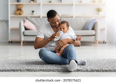 Childcare Concept. Loving African American Father Giving Bottle With Water To Baby Son At Home, Black Young Dad Spending Time With His Cute Infant Child, Sitting On Floor In Living Room, Copy Space