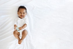 Childcare Concept. Portrait Of Adorable Little Black Baby Lying On Bed With Teether In Hand, Top View Shot Of Cute African American Infant Boy Or Girl Wearing Bodysuit Smiling At Camera, Copy Space