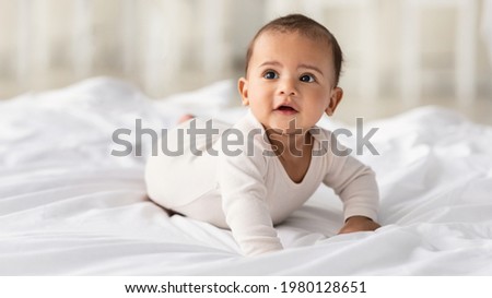 Childcare Concept. Portrait of cute little African American baby wearing bodysuit lying on white beedsheets at home. Black infant child crawling on bed in the bedroom. Selective focus, free copy space