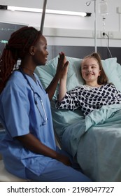 Childcare healthcare facility staff high fiving ill kid under medical treatment. Nurse doing high five gesture with sick girl resting in hospital pediatric ward patient bed.