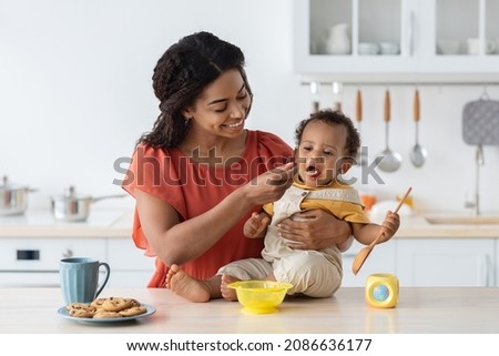 Childcare. Loving Black Mom Feeding Her Cute Baby Son From Spoon In Kitchen, Caring Mother Giving Porridge Or Mash Fruit Puree To Adorable Infant Child At Home, Preparing Healhy Food For Kid