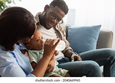 Childcare And Nursery. Happy Loving African American Parents Feeding Their Baby Boy From Bottle, Cute Black Toddler Drinking Water Or Milk. Smiling Family Sitting On Couch, Spending Time With Child