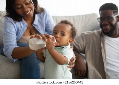 Childcare And Nursery. Happy Loving African American Parents Feeding Their Baby Boy From Bottle, Thirsty Cute Black Toddler Drinking Water. Smiling Family Sitting On Couch, Spending Time With Kid