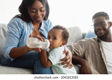 Childcare. Portrait Of Happy Loving Black Mother Feeding Her Baby Boy From Bottle, Cute Black Toddler Drinking Water Or Milk. Smiling Afro Parents Sitting On Couch, Spending Time With Their Child