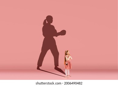 Childhood And Dream About Big And Famous Future. Conceptual Image With Girl And Shadow Of Strong Female Taekwondo Practitioner On Coral Pink Background. Sport, Dreams, Imagination, Education Concept.