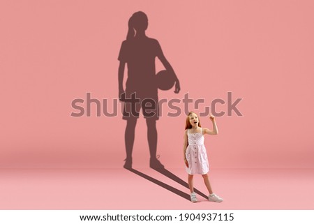 Childhood and dream about big and famous future. Conceptual image with girl and drawned shadow of basketball, soccer female player on coral pink background. Childhood, dreams, education concept.