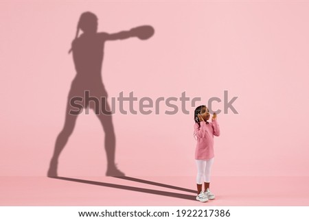 Childhood and dream about big and famous future. Conceptual image with girl and drawned shadow of female boxer on coral pink background. Childhood, dreams, imagination, education concept.