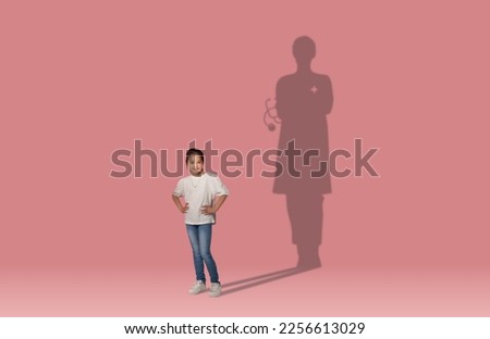 Childhood dreams concept. Cool cheerful school aged asian girl dreaming to become a doctor, shadow of female doc behind her, kid posing over pink wall studio background, collage, copy space