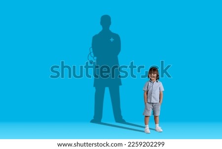 Childhood dreams concept. Cute handsome little boy dreaming to become a doctor, shadow of male doc behind kid posing over blue wall studio background, collage, copy space