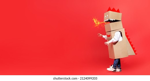 Childhood dreams. Funny little boy plays a cardboard dragon breathing fire and walking on a red background. Fantasy, imagination. Full-length studio portrait. Copy space.