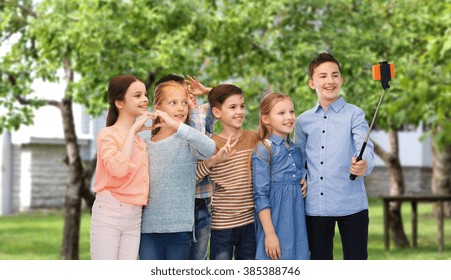 Childhood, Friendship, Technology And People Concept - Happy Children Talking Picture By Smartphone On Selfie Stick Over Private House Backyard Background